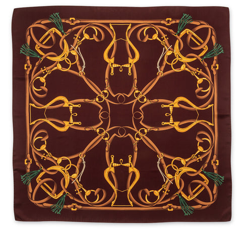 A square silk scarf featuring an intricate equestrian design with golden horse bits and reins on a dark brown background, finished with green tassels in the corners.