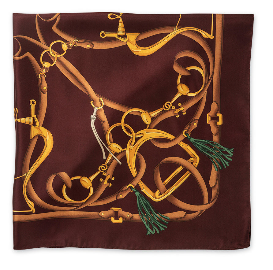 A square silk scarf featuring an intricate equestrian design of golden horse bits and reins over a deep burgundy background, finished with green tassels at the corners.